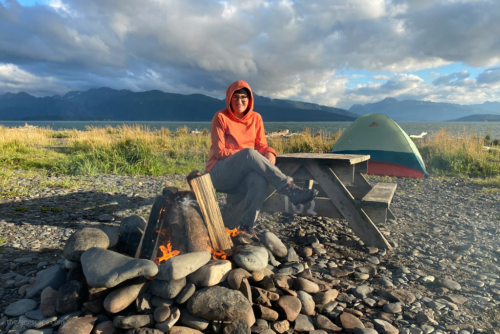 Agnes on Homer RV campground sitting next to campfire with sea and mountains in the backdrop.
