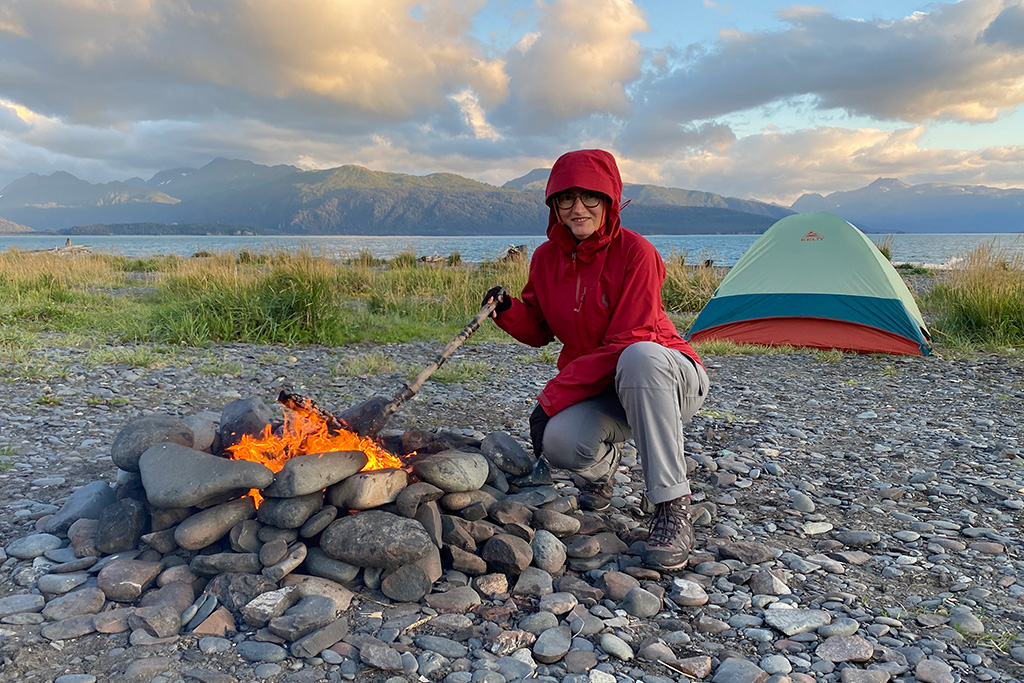 What to wear in Alaska? Agnes wearing red windstopper Black Diamond jacket, hiking pants and hiking boots on the camping in Homer with water and mountains in backdrop.