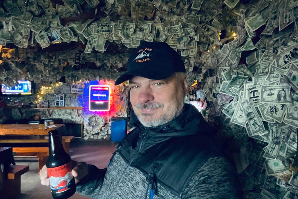 Smiling, Chris drinks an Alaskan beer in Salty Dwang Saloon with thousands of paper dollars on the walls.