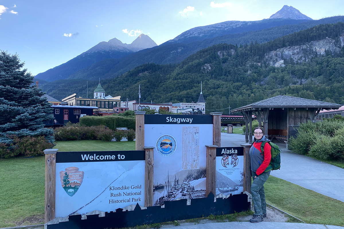 Agnes Stabinska, author, with skagway sign with mountains in the backdrop.