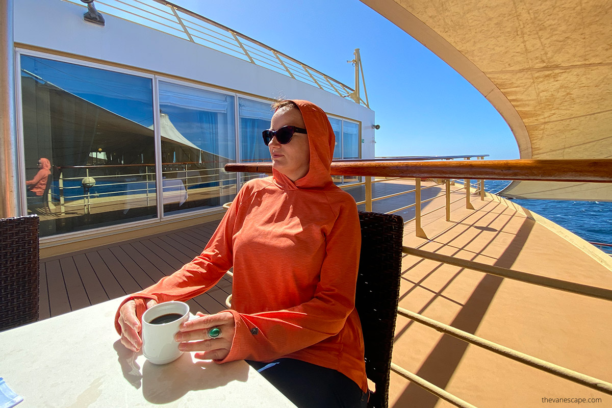 Agnes drinking coffee at ship during cruise, wearing orange sun protection t-shirt with a hood and sunglasses.