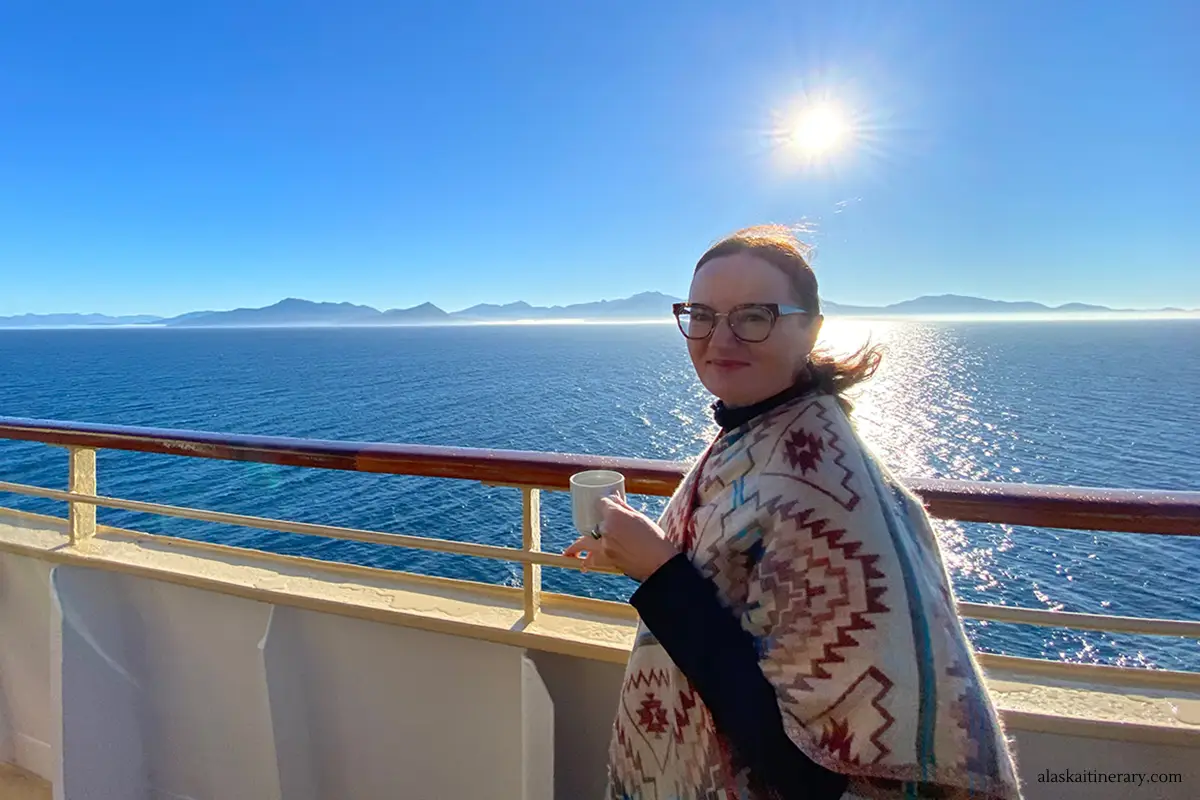 Agnes Stabinska, the author and owner of the Alaska Itinerary blog, is stanging on a cruise ship to Alaska and drinking a coffee with mountains and sun in the bacdrop.