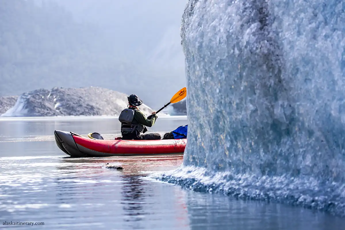 A kayaker enjoys glacier kayaking in Alaska, paddling a red kayak near the towering, icy face of a glacier, with a misty mountain backdrop.
