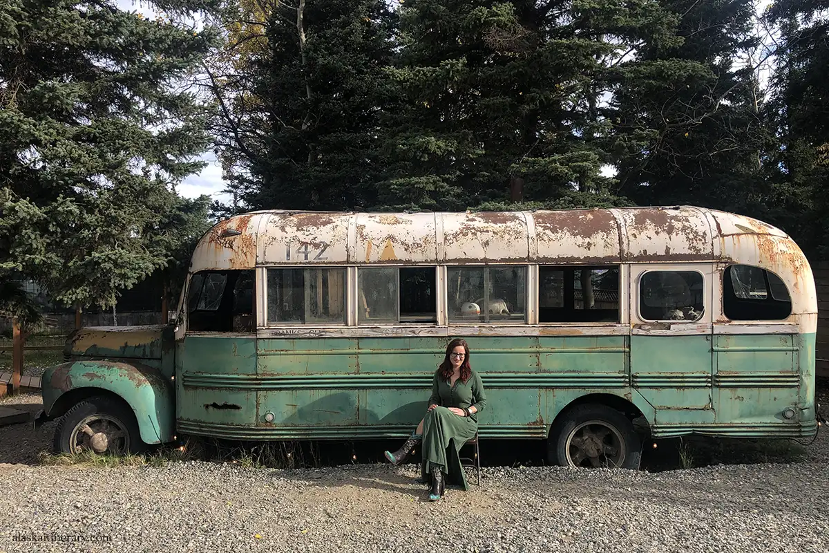 Agnes in green long dress sitting at the front of a replica of Magic Bus 142 in Healy from the movie Into the wild.