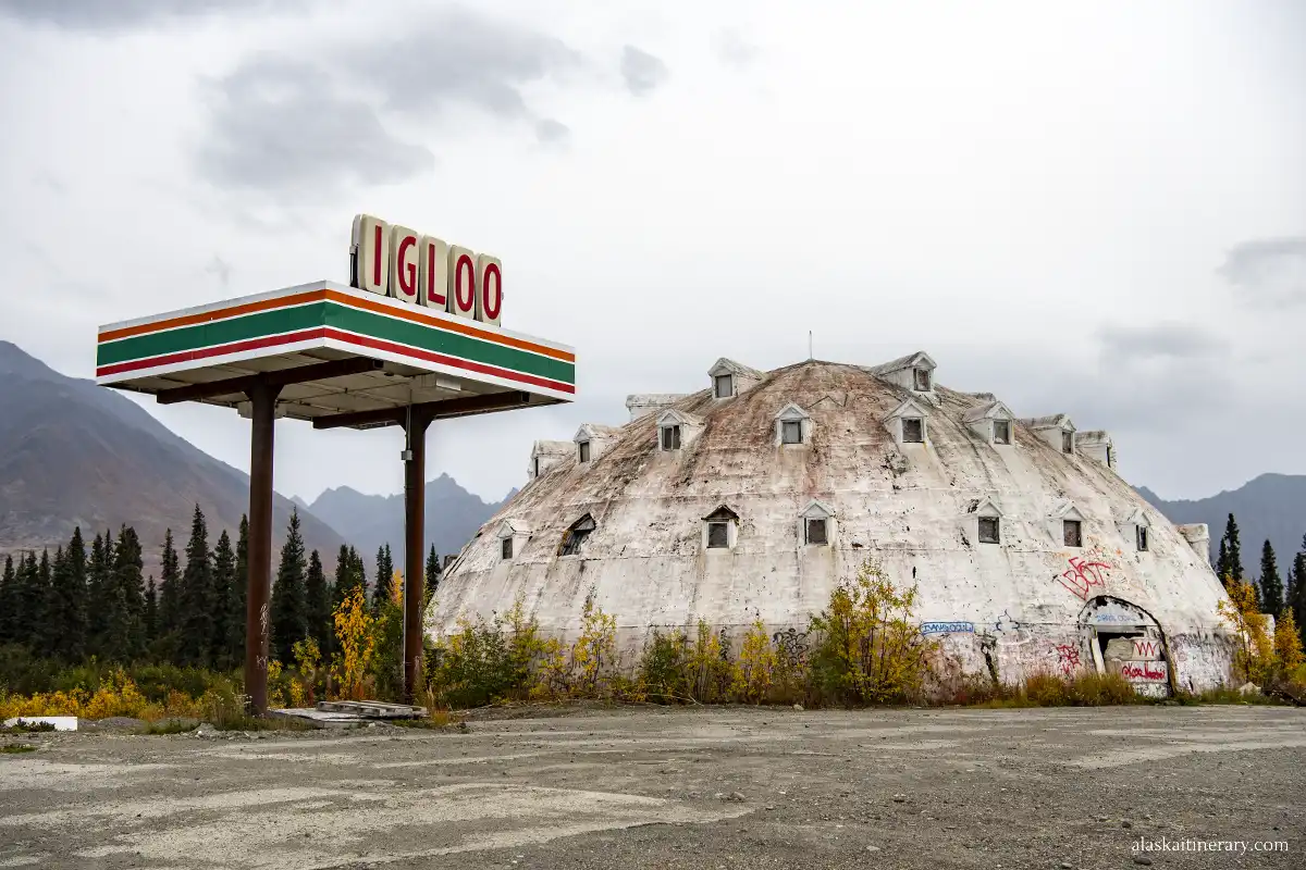 An abandoned, graffiti-covered building shaped like a large igloo stands beside a dilapidated gas station sign that reads 'IGLOO.' The scene is set in a desolate area with overgrown vegetation, under a cloudy sky.