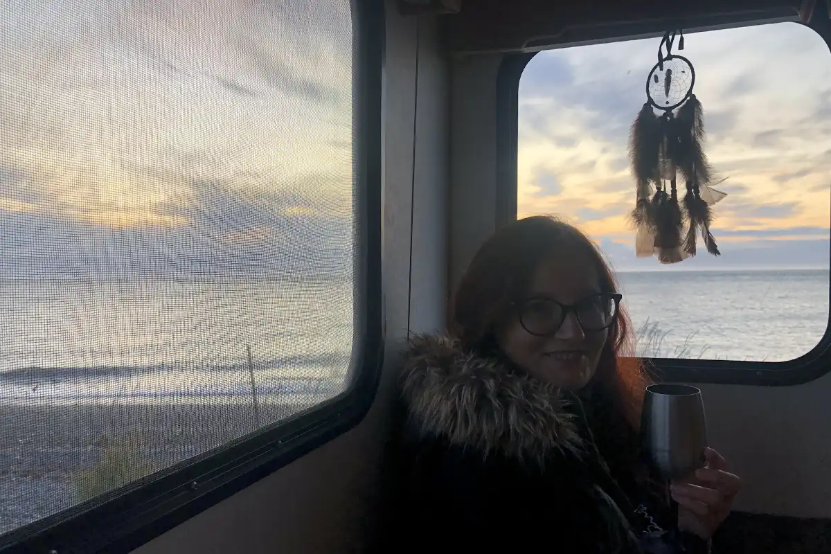 Agnes enjoying a cozy moment inside an RV, sipping from a wine glass with a dreamcatcher hanging by the window, overlooking the Alaskan sunset.
