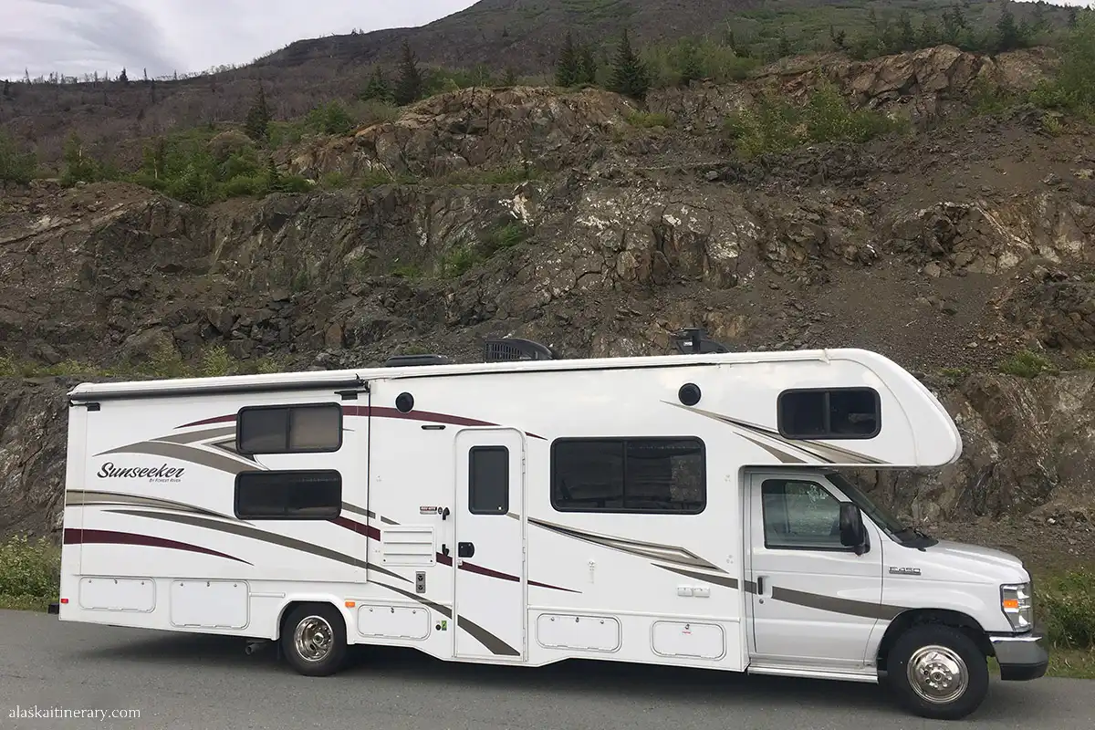 Class C motorhome named 'Sunseeker' parked on a gravel lot in Alaska, with rugged hills in the background.