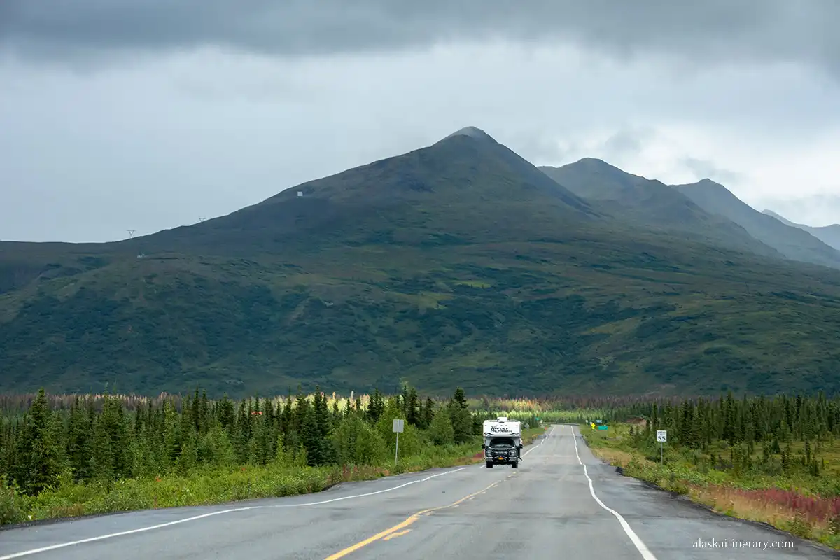 An RV driving on a paved road through a vast Alaskan landscape with rolling green hills and a mountain in the background. 