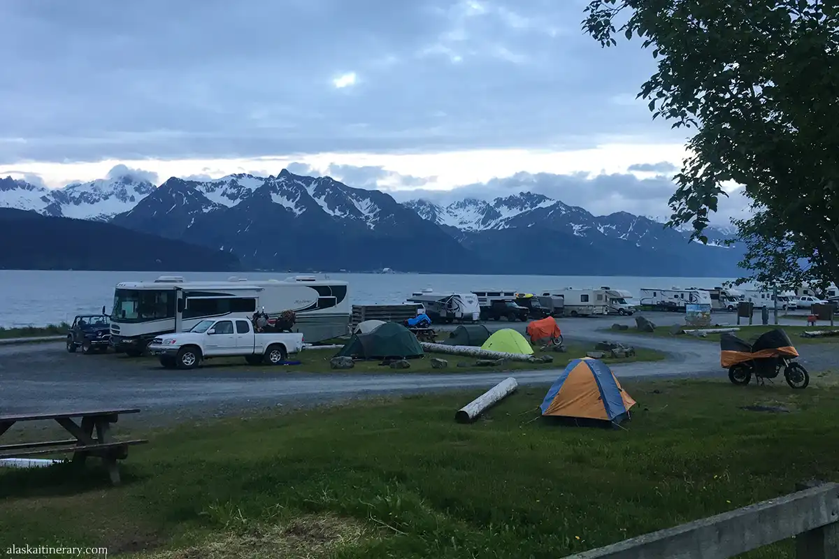 Scenic RV campground in Alaska with RVs, tents, a picnic area, and travelers enjoying the oceanfront view, set against a backdrop of towering snow-capped mountains.