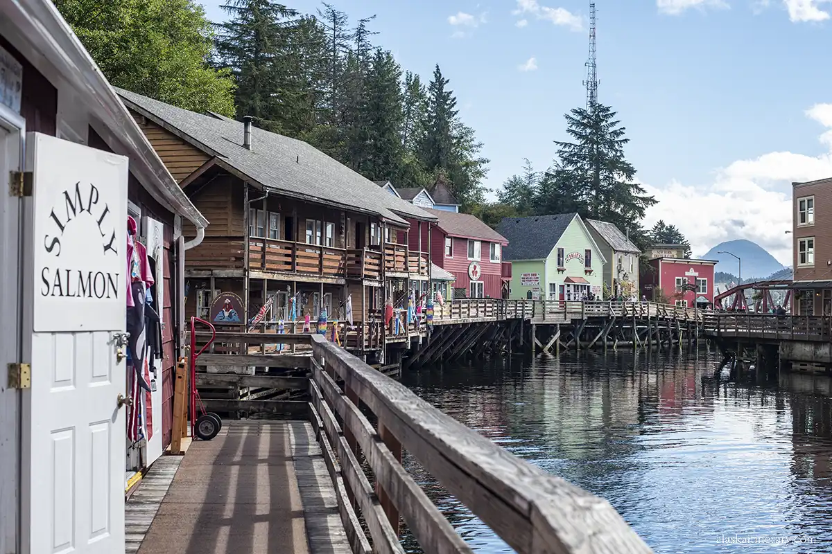 Creek Street  in Ketchikan with houses on stilts in the water,  in beautiful green, brown and pink pastel colors, and a wooden sidewalk.