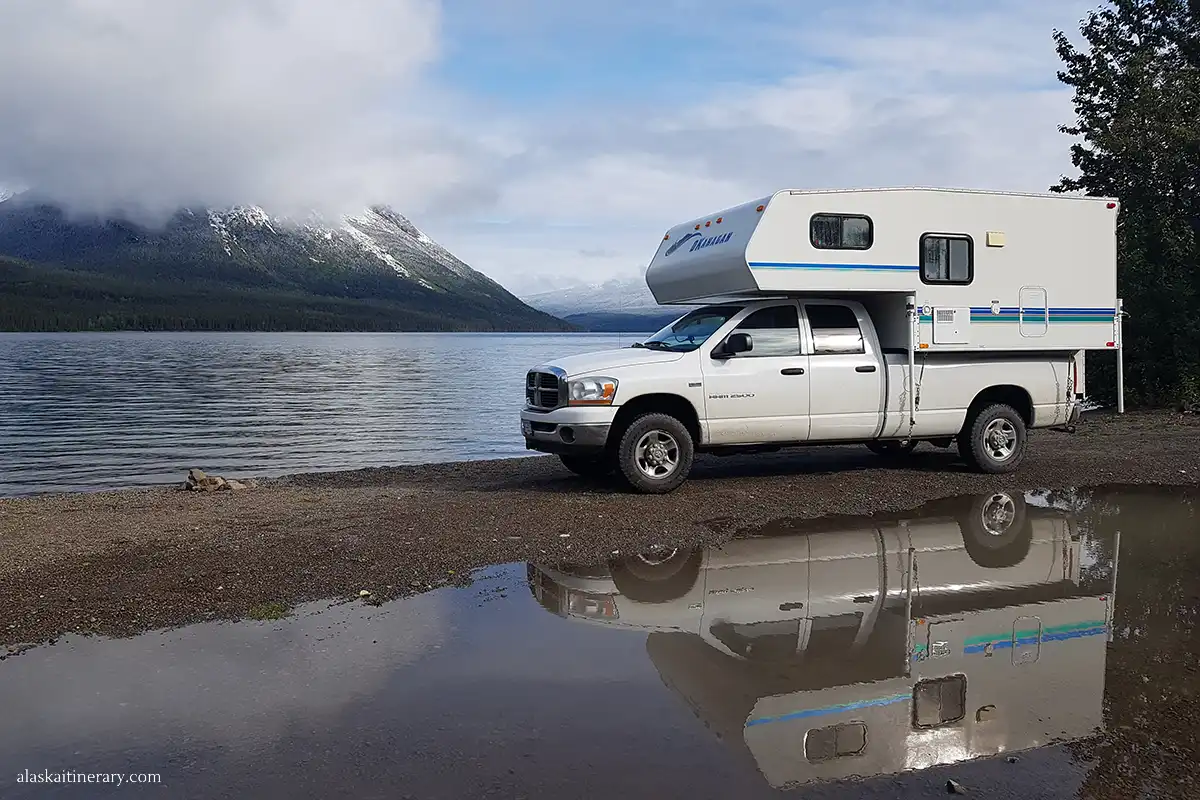 Our RV parked by a serene lake with a mountain range in the background and its reflection on the water's surface in Alaska