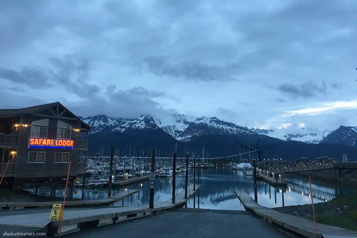 Dusk settles over Seward Harbor, Alaska, with the Safari Lodge overlooking a marina filled with boats, set against a stunning backdrop of snow-capped mountains reflecting the fading light.