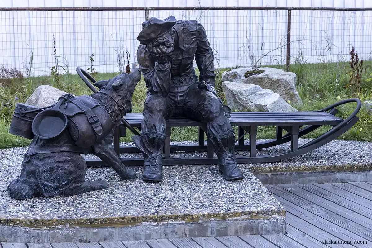 The image shows a bronze sculpture of a prospector and his pack dog, representing the Gold Rush era in Skagway, Alaska, with the dog eagerly looking up at the seated man, set against a rugged outdoor backdrop.