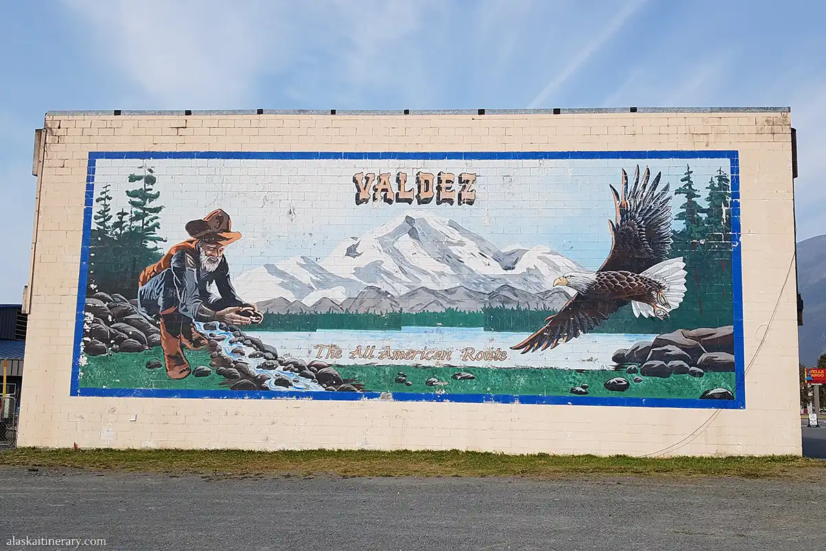 Mural in Valdez, Alaska, depicting a gold prospector and a soaring bald eagle, with the majestic Alaskan mountainscape in the background, symbolizing the 'All American Route'