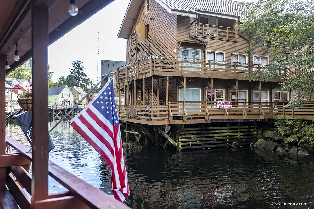 An American flag hangs over the creek; a large wooden house on stilts is in the background.
