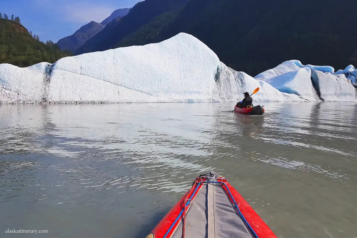 Paddling close to the massive, white wall of an Alaska glacier in a red kayak, with clear blue skies and mountains in the distance.