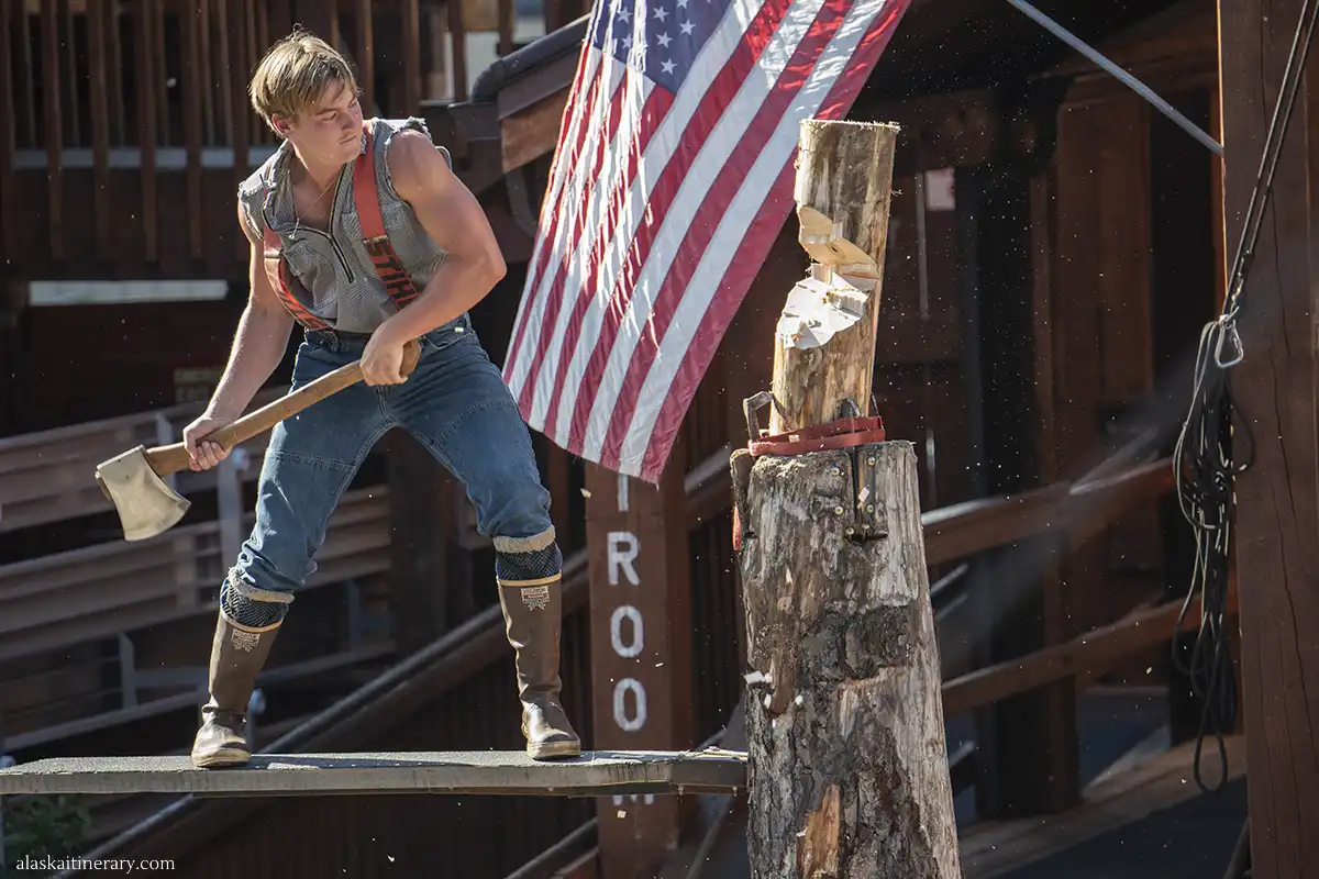 A young athlete with an ax in his hand cuts a tree trunk during a lumberjack show in Ketchikan. An American flag flies in the background.