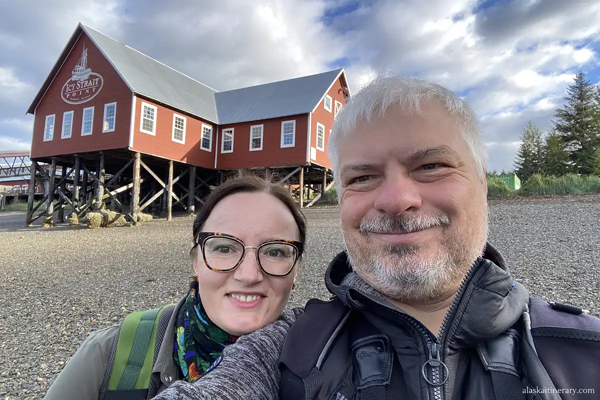 Agnes and Chris in Icy Strait Point cruise port with a brown wooden buildling of Hoonah Cannery in the backdrop.