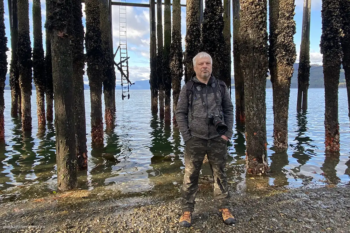 Chris stands on the beach under the pier at Icy Strait Point with the wooden pier posts in the background.