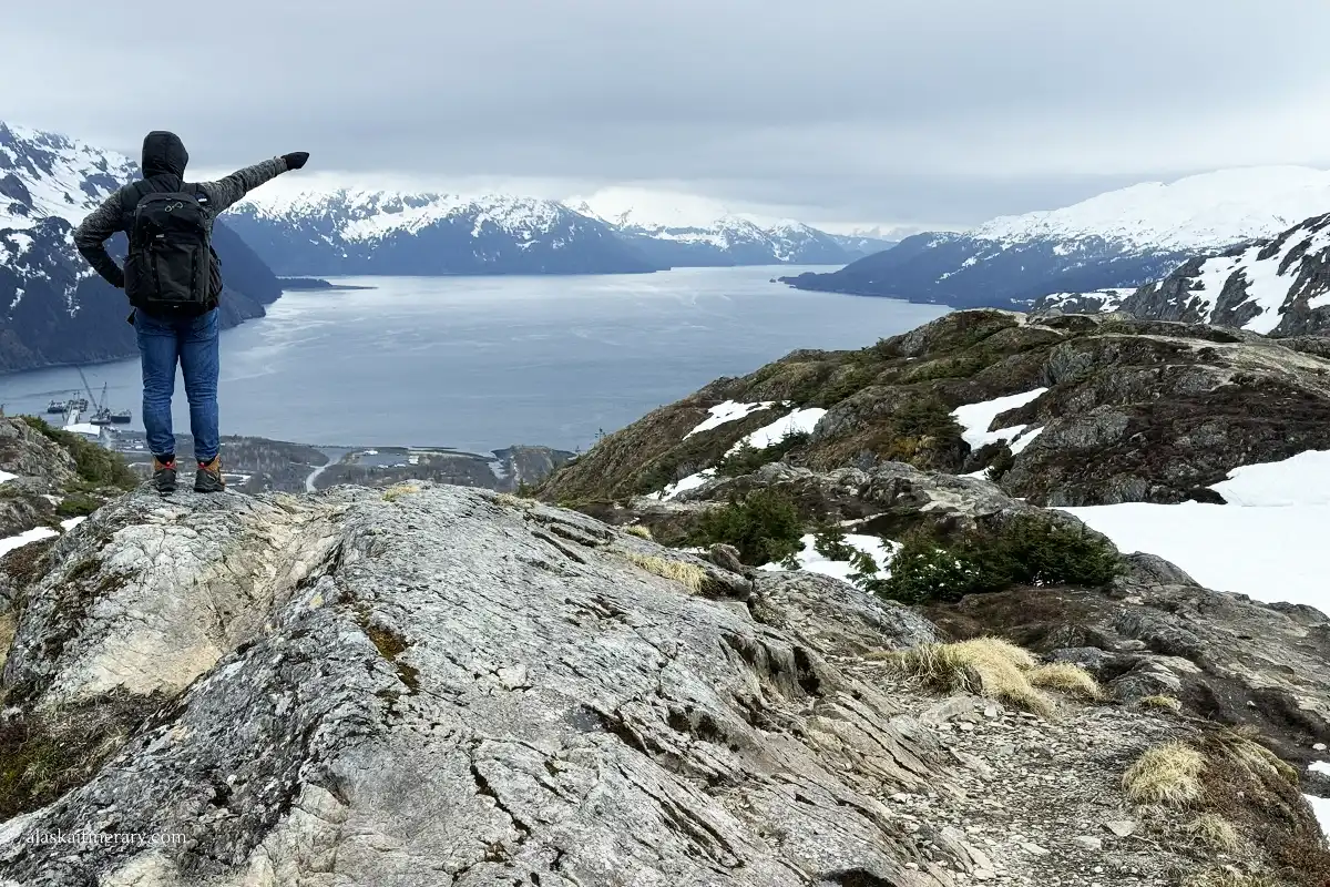 Chris stands on a rock after a steep uphill climb of the Portage Pass, with a panoramic view of Whittier, the bay, and the mountains; Chris is in a warm hoodie and a backpack, and his hand is pointing at Whittier.