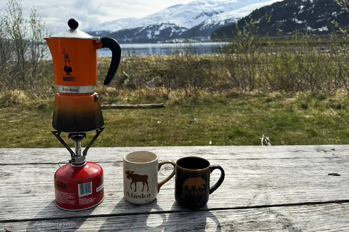 camping in Whittier: morning coffee with the mountains view: two mugs with coffee and orage coffee maker at picnic table.