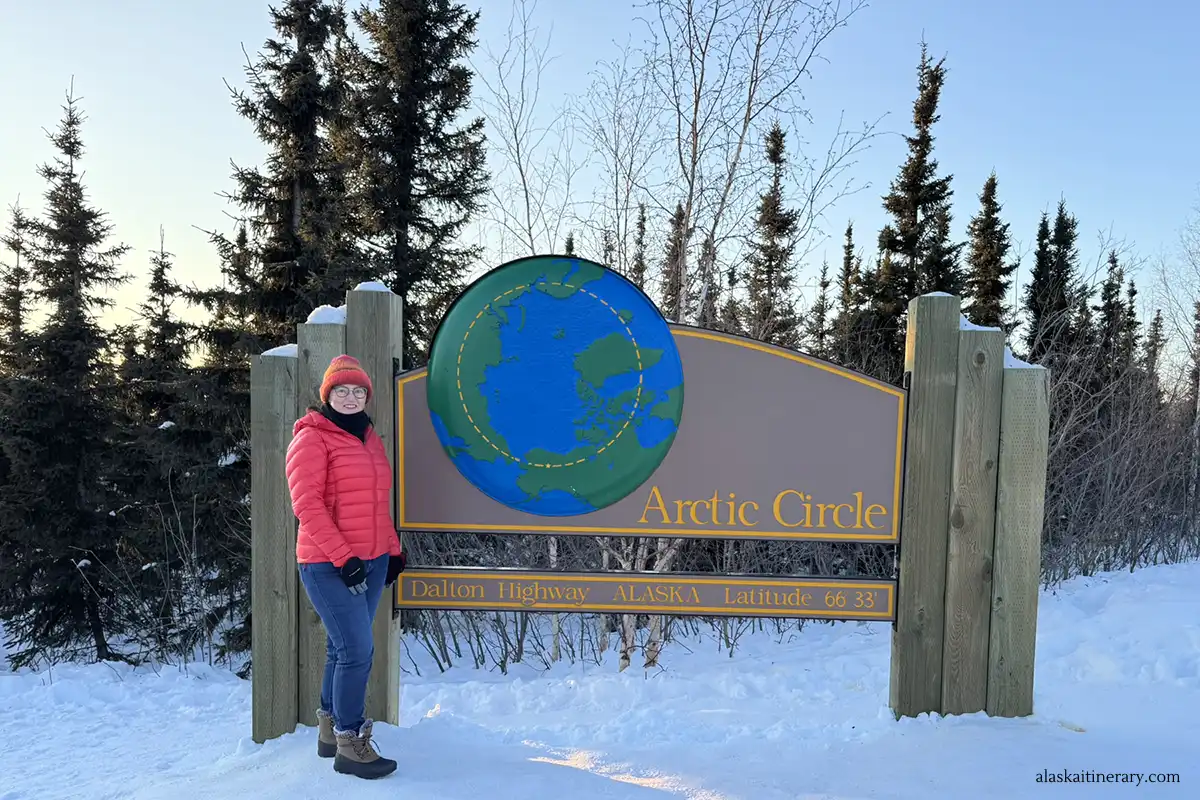Agnes Stabinska, the author, stands at the Arctic Circle sign in the winter scenery with the setting sun in the background.